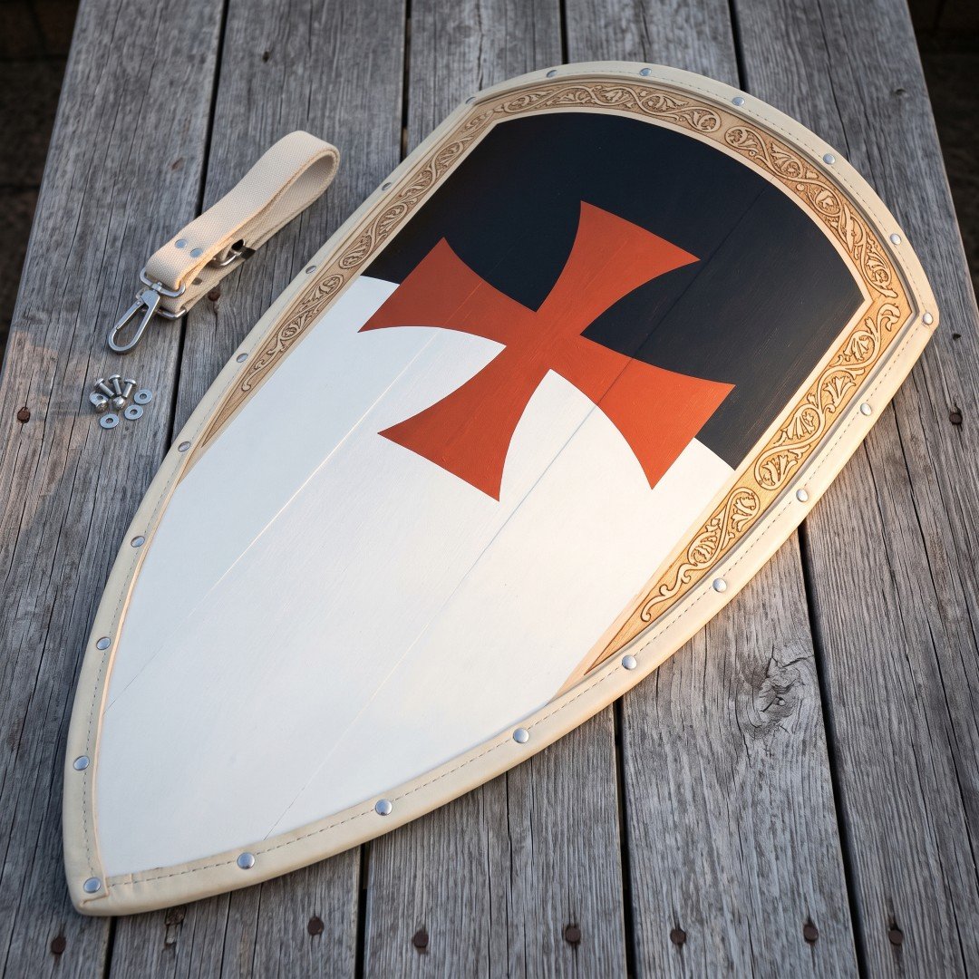 Templar kite shield of planked wood with a black and white field, a red cross pattee and an engraved gilt border strip, Templar kite shield of planked wood with a black and white field, a red cross pattee and an engraved gilt border strip, on grey decking beside a leather guige.