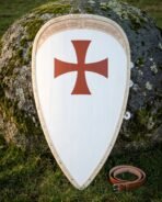 Templar shield of kite form in planked wood, a red cross pattee on a white field with a knotwork border band, leaning on a mossy boulder beside a leather belt.