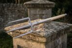 Three prong steel trident on a pale ash haft laid across a weathered stone pillar with a dry stone wall behind