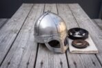 Three quarter view of a polished spectacle helmet with riveted brow band on weathered boards with liner and folded cloth