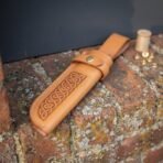 Tooled leather knife sheath with interlaced knotwork face and wide belt loop resting at an angle on a lichen covered brick wall