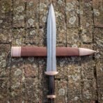 Top down view of a Damascus gladius short sword with bronze fittings lying across a bronze tipped leather scabbard on brick