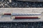 Top down view of a flamberge sword with wavy blade and brass fittings laid on driftwood above its red brown leather scabbard