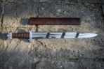 Top view of an arming sword with brown leather grip and steel cross laid next to a brown leather scabbard on stone