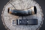 Top view of a dark ox drinking horn with steel bands above a raven engraved black leather belt and steel buckle
