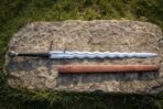 Top view of a full length steel flamberge sword with wavy blade and its brown leather scabbard on a flat stone