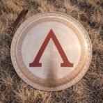 Top view of a Spartan hoplite shield with red lambda, Greek key border and studded rim lying on dry grass beside a leather strap