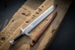 Triple fullered Viking short sword with ribbed black grip and downswept guard beside its brown leather scabbard