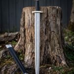 Triquetra damascus sword with a pattern welded Viking blade, three lobed pommel and black leather grip standing against a weathered tree stump