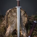 Triquetra Viking sword with a five lobed steel pommel and brown leather grip standing on a cut oak stump, its Damascus blade beside a brown leather scabbard