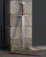 Triskele sword with a triple spiral wheel pommel and tan leather grip standing against a granite gatepost above its tooled tan leather scabbard with belt loops