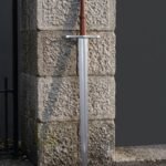Triskele sword with a triple spiral wheel pommel and tan leather grip standing against a granite gatepost above its tooled tan leather scabbard with belt loops