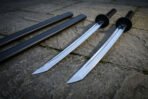 Twin ninjato set seen from the points on stone paving, showing the angled chisel style tips, mirror polished blades and the two black scabbards lying alongside.