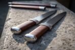 Twin seax knives with engraved bolsters and wrapped leather grips seen from the pommel end on a stone wall top