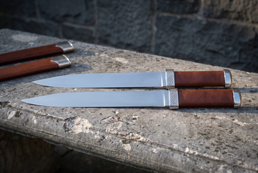 Twin seax knives in polished 1095 steel with brown leather grips and knotwork engraved steel bolsters lying on a stone ledge beside two brown leather sheaths