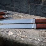 Twin seax knives in polished 1095 steel with brown leather grips and knotwork engraved steel bolsters lying on a stone ledge beside two brown leather sheaths