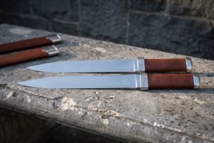 Twin seax knives in polished 1095 steel with brown leather grips and knotwork engraved steel bolsters lying on a stone ledge beside two brown leather sheaths