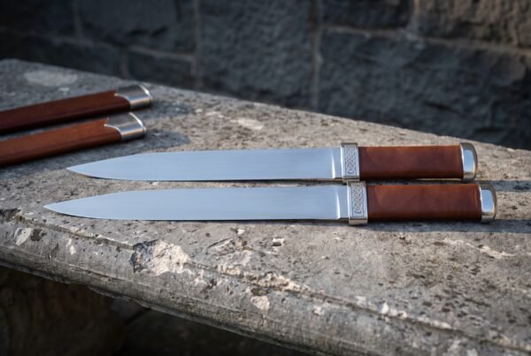 Twin seax knives in polished 1095 steel with brown leather grips and knotwork engraved steel bolsters lying on a stone ledge beside two brown leather sheaths