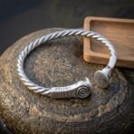 Twisted rope arm ring in silver finish with spiral terminals and rune engraved ends laid on a granite boulder, oak tray behind