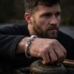 Twisted silver finish arm ring with spiral terminals and engraved runes worn on a bearded man's wrist as he leans on a rock