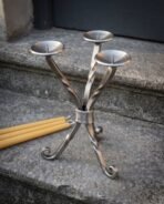 Twisted steel three branch candle holder with dished drip pans and scrolled feet on a stone step beside beeswax candles