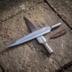 Two carbon steel spearheads with engraved sockets laid across each other on a stone slab with a leather blade cover