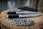 Two chain knives with black grips and ring pommels on a sawn log by the water, chains coiled and sheaths behind