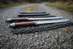 Two chain swords with oxblood grips and steel ring pommels lying on gravel, chains coiled beneath and scabbards behind