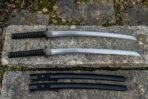 Two curved swords with black cord wrapped handles laid above two black scabbards on a lichen covered stone slab