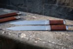 Two seax knives with leather grips laid parallel on a stone ledge, full length side view with both sheaths in the back