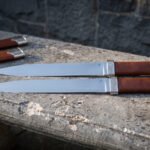 Two seax knives with leather grips laid parallel on a stone ledge, full length side view with both sheaths in the back