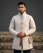 Undyed linen padded gambeson with tall collar and bow tied cord lacing worn with a brown leather belt by a dark haired man