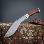 Valknut damascus kukri with rosewood scales and a brass bolster resting on a lichen covered brick wall beside its brown leather sheath and two companion blades.