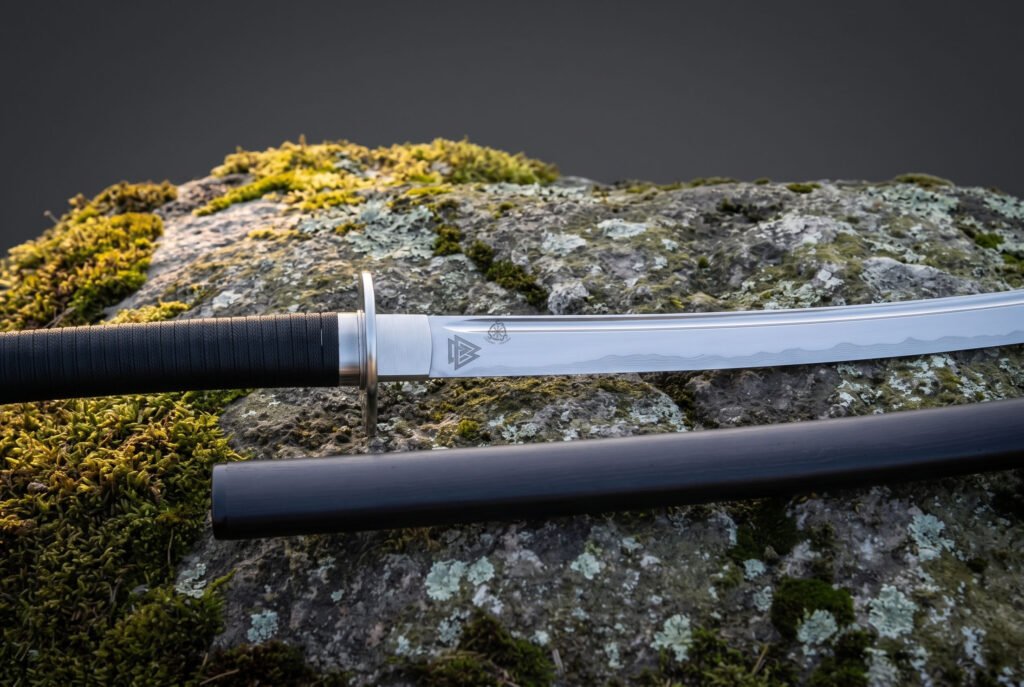 Valknut etched 1095 steel blade shown close up above the tsuba and black cord tsuka of a Viking katana on a lichen covered boulder