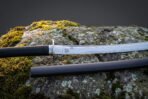 Valknut etched 1095 steel blade shown close up above the tsuba and black cord tsuka of a Viking katana on a lichen covered boulder