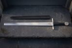 Valknut etched arming sword in satin 1095 steel with a plain square bar guard, black leather grip and flat disc pommel laid on dark slate beside its black scabbard