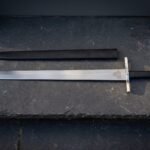 Valknut etched arming sword in satin 1095 steel with a plain square bar guard, black leather grip and flat disc pommel laid on dark slate beside its black scabbard