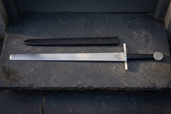Valknut etched arming sword in satin 1095 steel with a plain square bar guard, black leather grip and flat disc pommel laid on dark slate beside its black scabbard