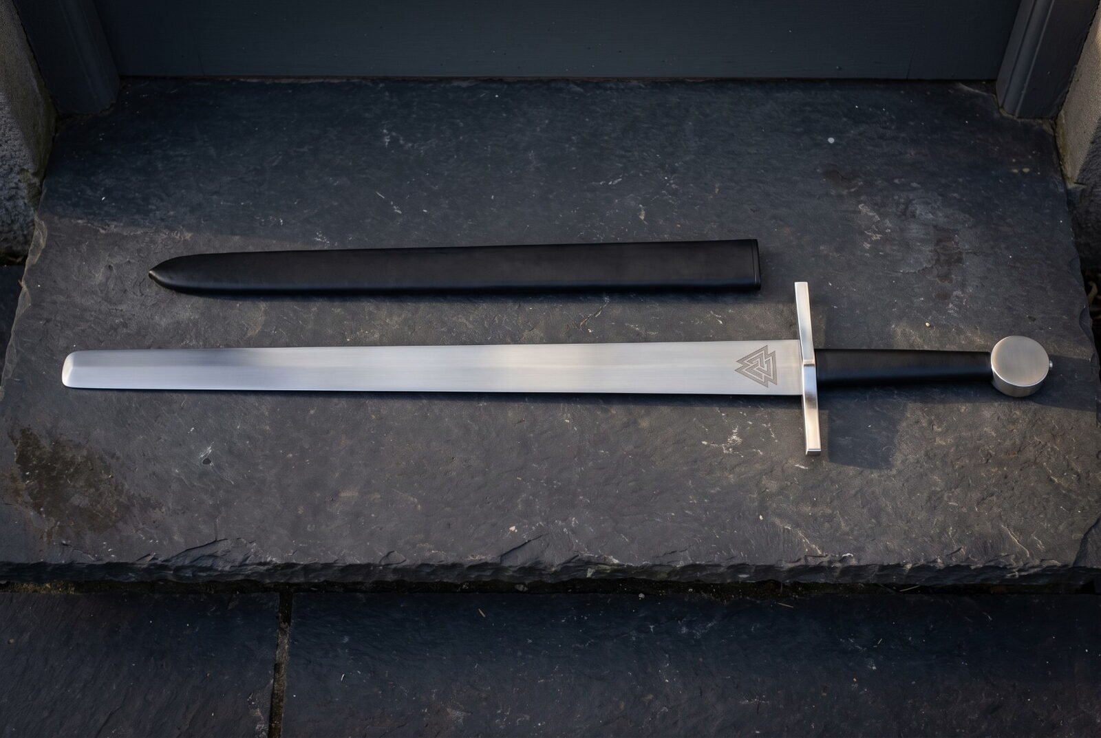 Valknut etched arming sword in satin 1095 steel with a plain square bar guard, black leather grip and flat disc pommel l Valknut etched arming sword in satin 1095 steel with a plain square bar guard, black leather grip and flat disc pommel laid on dark slate beside its black scabbard