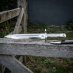 Valknut etched Viking bayonet in polished 1095 steel with a ribbed steel grip and muzzle ring guard resting on a weathered timber gate rail in a green field