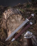 Valknut hilt viking sword laid across a mossy tree stump with its brown leather scabbard, showing the pattern welded fuller, curved guard and engraved Vegvisir mark