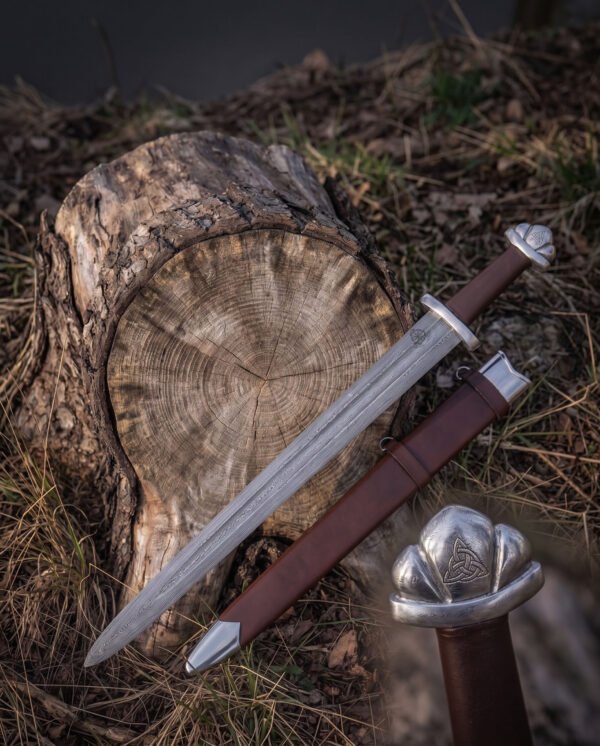 Valknut hilt viking sword laid across a mossy tree stump with its brown leather scabbard, showing the pattern welded fuller, curved guard and engraved Vegvisir mark