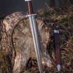 Valknut hilt viking sword propped against a cut tree stump in woodland, the five lobed pommel, brown leather grip and Damascus blade beside a brown leather scabbard