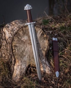 Valknut hilt viking sword propped against a cut tree stump in woodland, the five lobed pommel, brown leather grip and Damascus blade beside a brown leather scabbard