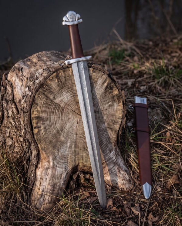 Valknut hilt viking sword propped against a cut tree stump in woodland, the five lobed pommel, brown leather grip and Damascus blade beside a brown leather scabbard