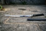 Valknut katana with three interlocked triangles etched at the blade base, a polished steel tsuba and black cord over white rayskin resting on grey stone paving