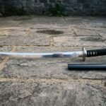 Valknut katana with three interlocked triangles etched at the blade base, a polished steel tsuba and black cord over white rayskin resting on grey stone paving
