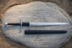 Valknut longsword in mirror polished 1095 steel laid across a pale sandstone boulder beside its black leather scabbard, showing the twin hooked guards and black grip.
