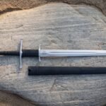 Valknut longsword in mirror polished 1095 steel laid across a pale sandstone boulder beside its black leather scabbard, showing the twin hooked guards and black grip.