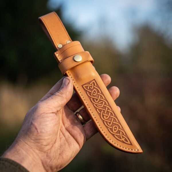 Vegetable tanned leather knife sheath with Celtic knot tooling and brass snap held upright in hand against autumn trees